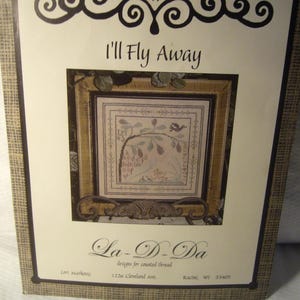 May include: A cross-stitch pattern kit titled "I'll Fly Away" by La-D-Da. The kit features a framed cross-stitch design with a bird, floral elements, and text. The packaging has a decorative border and the designer's name and address.