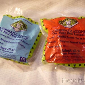 May include: Two sealed McDonald's Happy Meal toys featuring a Cabbage Patch Kids theme. One is blue, the other orange, both with text reading "Congratulations On Your New Arrival!" and the McDonald's logo.