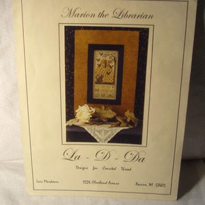 May include: A printed paper featuring a cross-stitch design titled "Marion the Librarian." The design is framed within a larger border, with the text "La - D - Da" below. The paper includes the designer's name and address.