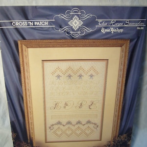 May include: Cross stitch pattern for a sampler titled "The Hope Sampler" by Emie Bishop. The pattern features an alphabet, the word "HOPE" and a border design. The pattern is framed in a gold frame and is on a blue background.