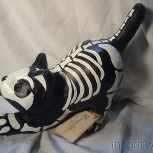 May include: A black cat figurine featuring a white painted skeleton design across its body. The cat is in a stretching pose with its tail raised. A small rectangular tan paper tag with the text "Bethany Lowe Designs" is attached to the cat.