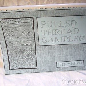 May include: A light blue book with the title "PULLED THREAD SAMPLER" and the author "Ilse Altherr". The book cover features a black and white pulled thread sampler design.