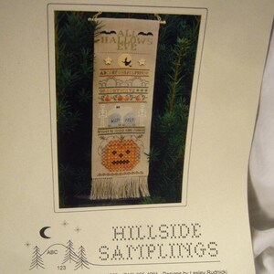 May include: A framed cross-stitch sampler with the text "ALL HALLOW'S EVE" at the top. The sampler features Halloween-themed motifs, including bats, stars, a pumpkin, and tombstones. The bottom of the sampler has a fringed edge.