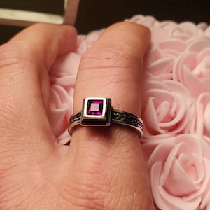 May include: A silver ring with a square, magenta-colored stone set in the center. The ring has a decorative band with a dark, textured pattern. The ring is on a finger, with a background of pink roses.