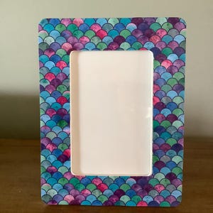 May include: Rectangular photo frame with a colorful mermaid scale pattern in shades of blue, purple, green, and pink. The frame has a white interior and is designed to hold a photograph. The frame is made of a sturdy material.