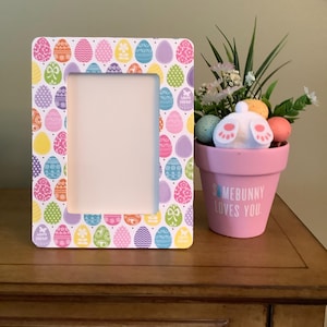 May include: A rectangular photo frame with a colorful Easter egg pattern. A pink flower pot with the text "SOMEBUNNY LOVES YOU" and a bunny tail decorates the scene. The frame is on a wooden surface.