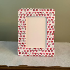 May include: A rectangular picture frame with a white background and a pattern of red and pink hearts. The frame has rounded corners and a white interior space for a photo. The frame is sitting on a white surface.