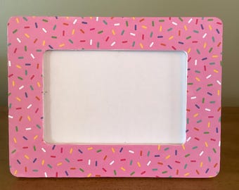 May include: A pink rectangular picture frame with a white interior. The frame is decorated with colorful sprinkles in various colors, including red, yellow, green, and blue. The frame is sitting on a wooden surface.