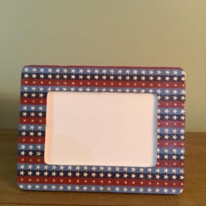May include: Rectangular photo frame with a patriotic design. The frame features alternating stripes of red, white, and blue, adorned with white stars. The frame is made of wood and has a white interior.