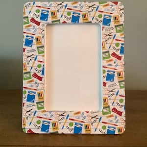 May include: A rectangular picture frame featuring a repeating pattern of school supplies including notebooks, pencils, scissors, paper clips, and green apples on a white background with blue horizontal lines and the text no more homework.