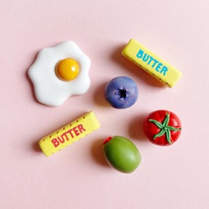 Six food-themed magnets on a soft pink background, a sunny-side-up egg, a blueberry, a green pimento olive, a bright red tomato with a green stem, and two sticks of butter with “butter” written on them; one in blue text and the other with red text.