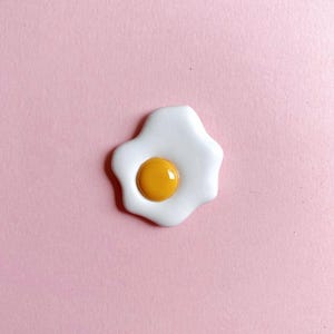 A sunny-side-up fried egg on a soft pink background. The egg yolk is glossy, which lends to its realism. A great gift for a food-lover/foodie/cook.
