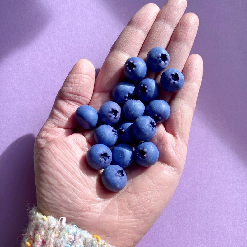 A hand with a palmful of realistic-looking polymer clay blueberries, set against a lilac background.