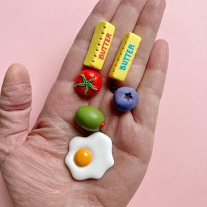 A hand holding six polymer clay food-themed magnets: a red tomato with green leaves and a green stem, a green olive with a red pimento center, a blueberry, a sunny-side-up egg, and 2 sticks of butter: one salted (red details) and one unsalted (blue details).