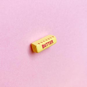 A pale yellow, polymer clay butter stick magnet with the word “butter” written in all caps with measurement marks along the side in red to represent salted butter. The ends of the butter stick have indentations to resemble the folded paper packaging.