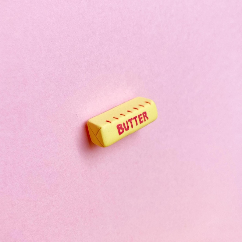 A pale yellow, polymer clay butter stick magnet with the word “butter” written in all caps with measurement marks along the side in red to represent salted butter. The ends of the butter stick have indentations to resemble the folded paper packaging.