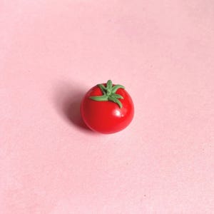 A mini red tomato polymer clay magnet with green leaves and stem. The tomato has a smooth, glossy surface and is set against a light pink background. A great gift for a food-lover/foodie/cook.