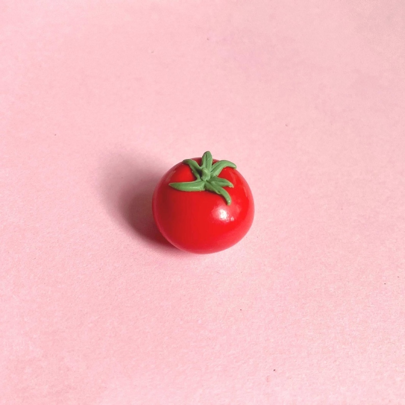 A mini red tomato polymer clay magnet with green leaves and stem. The tomato has a smooth, glossy surface and is set against a light pink background. A great gift for a food-lover/foodie/cook.