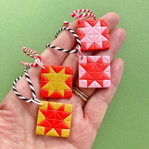 Peut inclure: Quatre ornements carrés avec des motifs d'étoiles rouges, roses et jaunes. Chacun est attaché à un cordon torsadé rouge, blanc et noir. Les ornements sont présentés sur un fond vert.