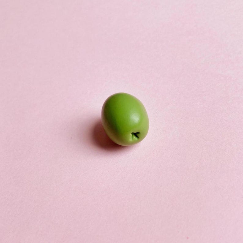 A green pimento olive, with a star impression on one end, sits on a soft pink background—a great gift for a cocktail-lover or martini-lover.