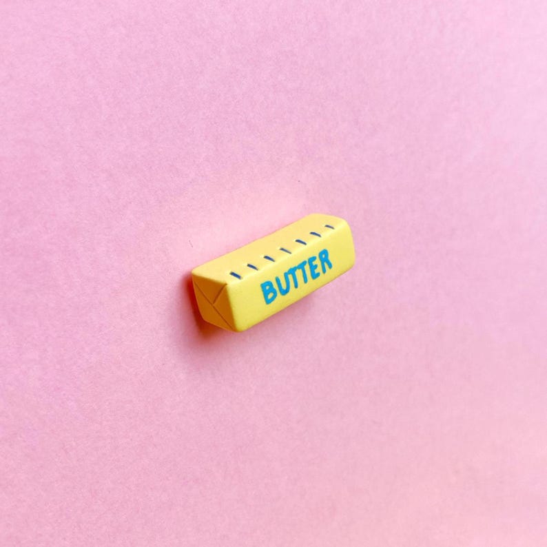 A pale yellow, polymer clay butter stick magnet with the word “butter” written in all caps with measurement marks along the side in blue to represent unsalted butter. The ends of the butter stick have indentations to resemble the folded paper packaging.