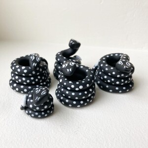 May include: Four black and white polka dot ceramic snake figurines. The snakes are coiled in a circle and have white eyes and markings. The figurines are different sizes.