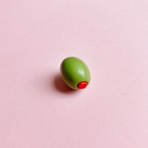 A green olive, stuffed with a red pimento, sits on a soft pink background—a great gift for a cocktail-lover or martini-lover.