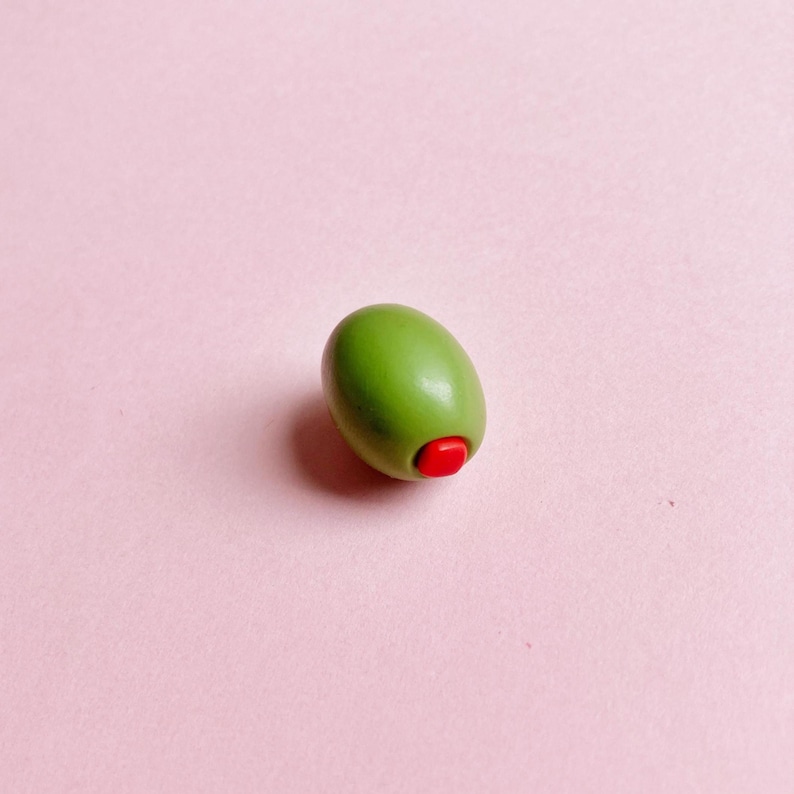 A green olive, stuffed with a red pimento, sits on a soft pink background—a great gift for a cocktail-lover or martini-lover.