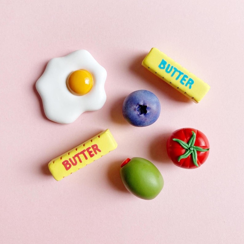 Six food-themed magnets on a soft pink background, a sunny-side-up egg, a blueberry, a green pimento olive, a bright red tomato with a green stem, and two sticks of butter with “butter” written on them; one in blue text and the other with red text.