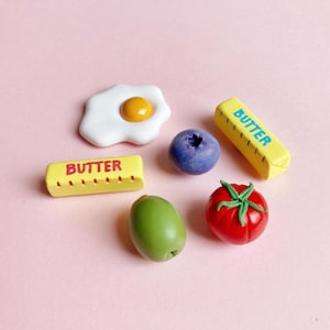 Six handmade polymer clay food-themed magnets on a pink background: a sunny-side-up egg, a green pimento olive, a bright red tomato with green leaves and stem, and two butter stick magnets labeled "butter" — one in blue text and one in red text, each with measurement marks on the sides.