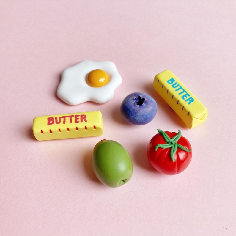 Six handmade polymer clay food-themed magnets on a pink background: a sunny-side-up egg, a green pimento olive, a bright red tomato with green leaves and stem, and two butter stick magnets labeled "butter" — one in blue text and one in red text, each with measurement marks on the sides.