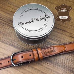 May include: A brown leather belt with a silver buckle and the name "Steward Wright" engraved on it. The belt is next to a silver tin with the same name on the lid. The words "Genuine Leather" are also visible.