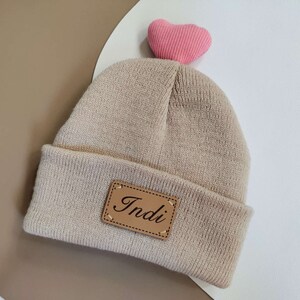 Puede incluir: Gorro de punto beige con un parche de cuero marrón que dice "Indi" y un corazón rosa en la parte superior.