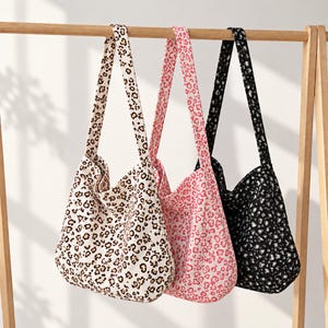 May include: Three hobo-style shoulder bags hanging from a wooden rack. Each bag features a distinct leopard print pattern in different colour combinations: one in beige and brown, one in pink and white, and one in black and white.