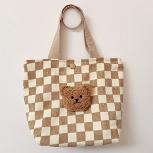 Könnte beinhalten: Eine kleine braune und weiße karierte Tragetasche mit einem braunen Teddybär-Applikationen. Die Tasche hat einen braunen Griff.