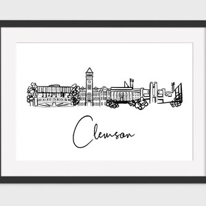 May include: Black framed print featuring a minimalist line drawing of the Clemson, South Carolina skyline. The word "Clemson" is written in cursive below the skyline. The artwork is in black and white.
