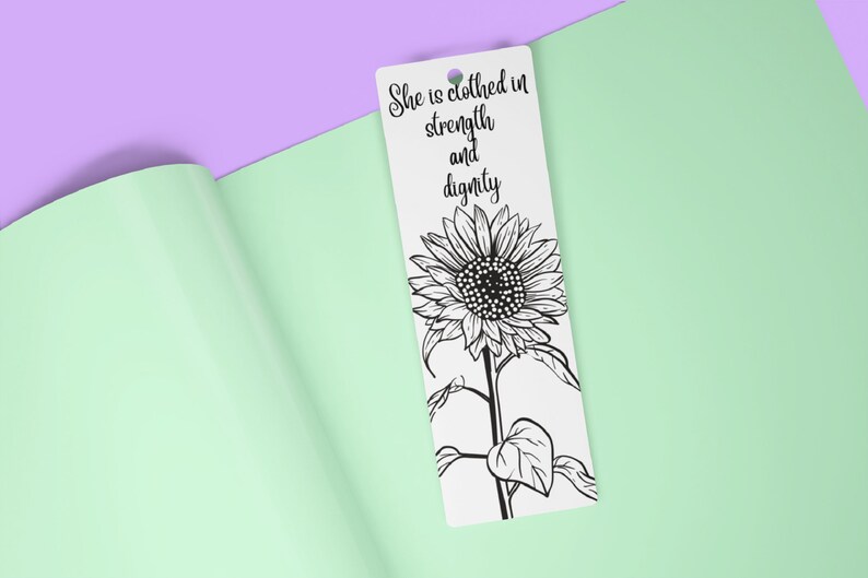 May include: A white bookmark featuring a black and white illustration of a sunflower. The text "She is clothed in strength and dignity" is printed on the bookmark.