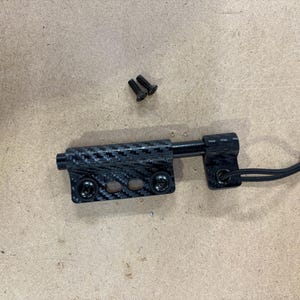 May include: A black and carbon fiber door latch with two small screws. The latch has a textured carbon fiber pattern and a black cord attached to one end. The latch is designed to secure a door or similar object.