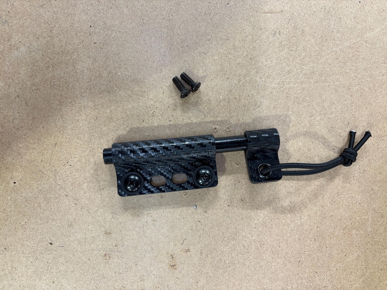 May include: A black and carbon fiber door latch with two small screws. The latch has a textured carbon fiber pattern and a black cord attached to one end. The latch is designed to secure a door or similar object.