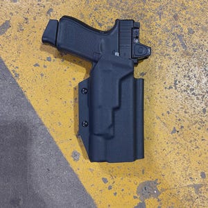 Puede incluir: Una pistola negra con mira de punto rojo está enfundada en una funda negra moldeada. El arma de fuego está colocada sobre una superficie amarilla y gris, probablemente de hormigón o asfalto. La funda está asegurada con tres tornillos negros.