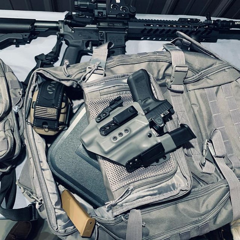 May include: A gray tactical backpack with a black handgun in a holster, a magazine, and ear protection. The backpack is open and the contents are visible.