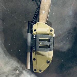 Puede incluir: Una funda de cuchillo verde y negra con un mango negro y un cord&oacute;n de paracord verde.