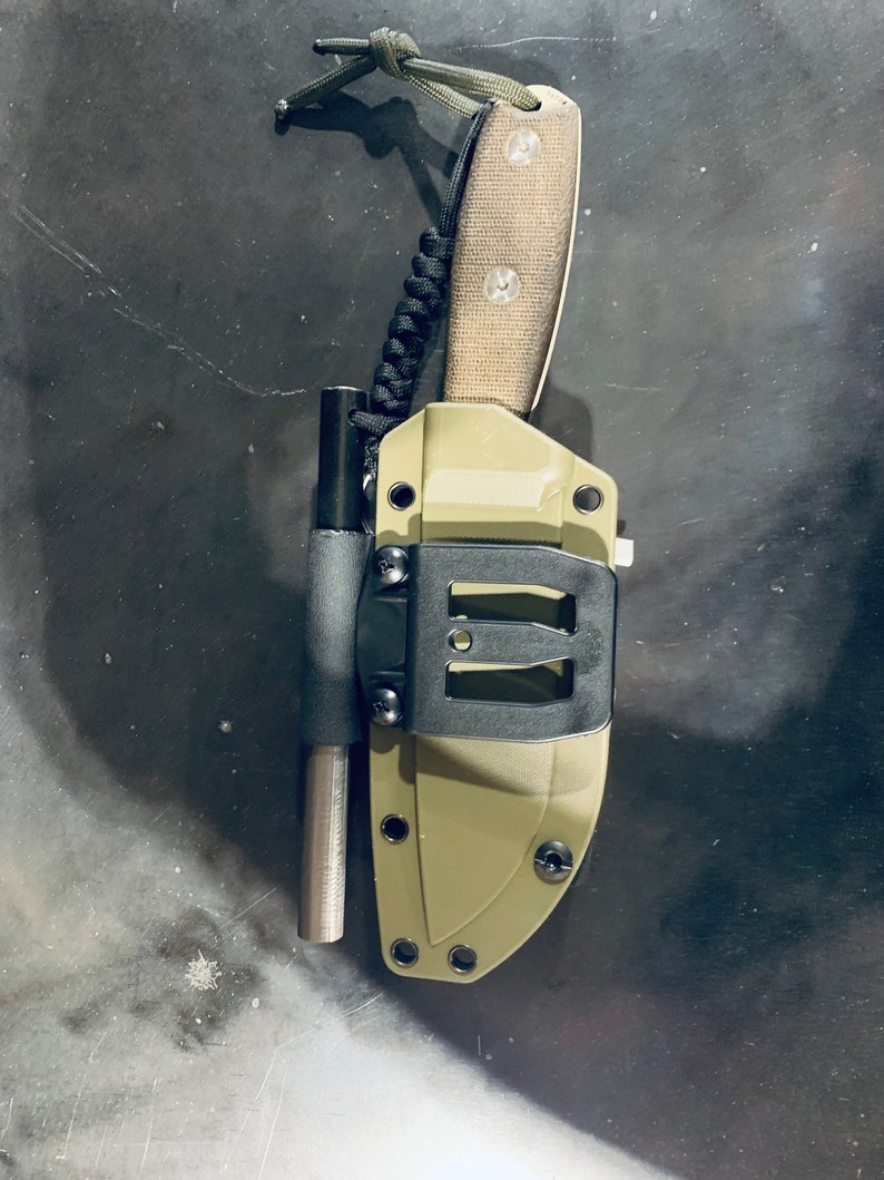 Puede incluir: Una funda de cuchillo verde y negra con un mango negro y un cord&oacute;n de paracord verde.