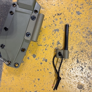 May include: An olive green knife sheath with black hardware and a matching ferro rod holder. A black ferro rod is next to the holder. The sheath has multiple eyelets and a lanyard. The items are on a yellow surface.