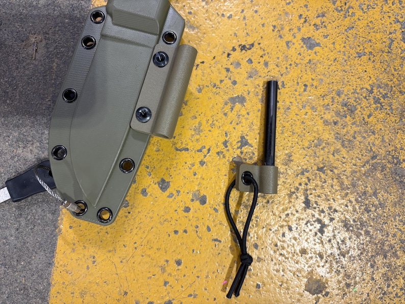 May include: An olive green knife sheath with black hardware and a matching ferro rod holder. A black ferro rod is next to the holder. The sheath has multiple eyelets and a lanyard. The items are on a yellow surface.