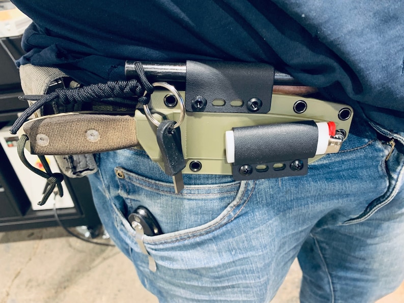 Puede incluir: Una persona que lleva pantalones vaqueros azules con una funda de cuchillo de cuero negro, un encendedor y un llavero sujetos a su cintur&oacute;n.