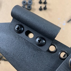 May include: Close-up of a black plastic holster with two black screws and four black eyelets. The holster has a textured surface.
