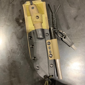 Puede incluir: Una funda de cuchillo negra con una funda de tela beige y un iniciador de fuego adjunto. La funda del cuchillo tiene una correa y un lazo para sujetar al cinturón.