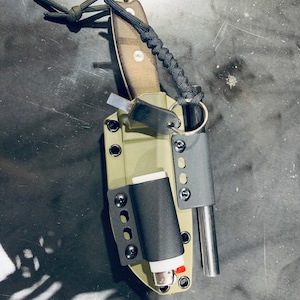 Puede incluir: Una funda de cuchillo verde con un encendedor negro, una varilla met&aacute;lica peque&ntilde;a y un cord&oacute;n de paracord negro atado.