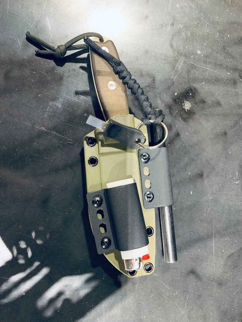 Puede incluir: Una funda de cuchillo verde con un encendedor negro, una varilla met&aacute;lica peque&ntilde;a y un cord&oacute;n de paracord negro atado.